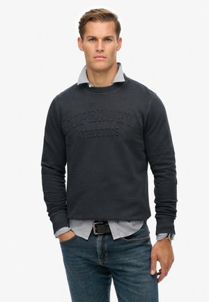 Granatowa bluza z wytłoczonym logiem "Superdry Athletics", z ściągaczami na rękawach i teksturowanym wewnętrznym kołnierzem, nałożona na jasnoniebieską koszulę w paski.