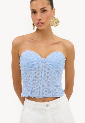 Femme portant un corset bustier en dentelle bleu clair et de grandes boucles d'oreilles dorées en forme de fleur, associées à un pantalon blanc sur un fond blanc.