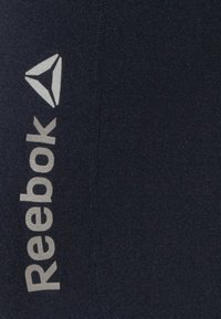 Reebok träningskläder i mörk marinblå. Har silverfärgad logotyp och triangulärt emblem. Materialet verkar ha en texturerad och elastisk kvalitet.