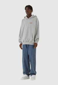 Grå hættetrøje med kengurulomme og subtilt logo, parret med løstsiddende blå jeans og hvide sneakers. Glat tekstur.