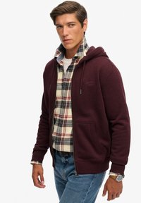 Haut à capuche bordeaux zippé avec cordon de serrage, poches avant, superposé sur une chemise à carreaux en crème, rouge et bleue. Jean en denim et montre au poignet.