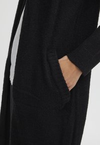 Schwarze Strickjacke mit strukturiertem Stoff, offenem Vorderteil und Seitentaschen. Die Ärmel sind gerippt für zusätzlichen Detailreichtum.