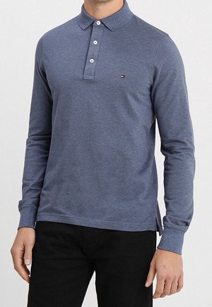 Mężczyzna w niebieskiej koszulce polo z długim rękawem, mający trzy guziki i noszący czarne spodnie, stoi na tle jednolitym.