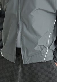 Veste coupe-vent grise avec une fermeture éclair, présentant des rayures blanches. Portée par-dessus un pantalon noir à motif à carreaux.