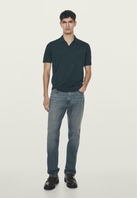 Ciemnozielona koszulka polo z krótkim kołnierzykiem, zestawiona z jasnoniebieskimi dżinsami i brązowymi butami, stojąca na neutralnym tle.