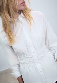 Donna bionda che indossa un vestito camicia bianco con dettaglio in vita cinte, in piedi contro uno sfondo neutro.