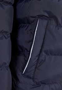 Marineblå pufferjakke-seksjon med quiltet tekstur, slankt design og en sidelomme med hvit pipingdetalj.