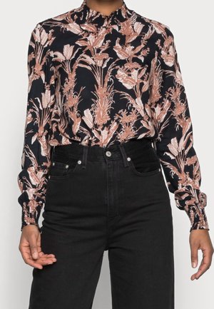 Chemisier à manches longues avec un motif floral rose et crème sur un fond sombre. Il présente un col montant et des manches froncées. Porté avec un jean noir.