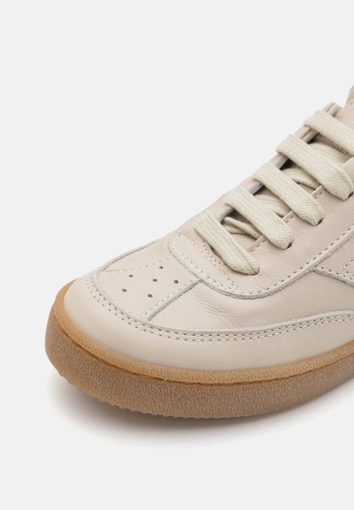 Beige lädersneaker med texturerad ovandel, rund tå och beige snören. Har perforeringar på tån och en gummisula.