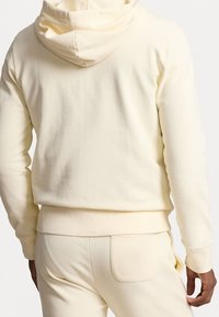 Tył osoby w kremowej bluzie z kapturem i pasujących dresach, ukazujący szczegóły kaptura i tylnej kieszeni.