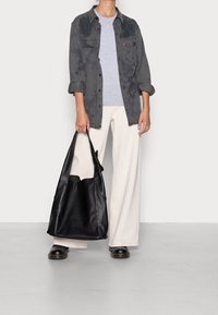 Sac cabas en cuir noir à la texture douce, doté de poignées larges et d'un design souple. Porté avec un pantalon large de couleur claire et un t-shirt gris.