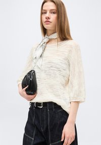 Jeune femme portant un pull léger en maille, une ceinture noire et un jean foncé, tenant une pochette noire avec un foulard blanc à motif autour du cou.