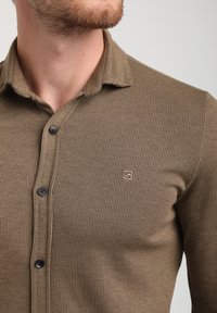 La maglietta a maniche lunghe a coste marrone presenta un design con bottoni, bottoni neri e un piccolo logo ricamato sul petto.