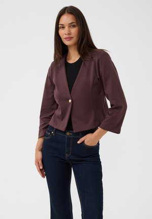 Femme aux cheveux raides et foncés portant un blazer court prune sur un top noir et un jean bleu foncé, une main dans la poche, sur fond blanc.