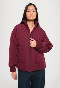 Wine-colored quilted jacket with a high collar, zip closure, and two front pockets. Features a relaxed fit and ribbed cuffs.