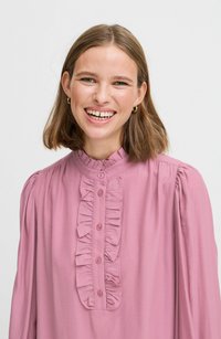 Rosa Bluse mit langen Ärmeln, Rüschendetail an der Vorderseite, Knopfverschluss und gerafften Schultern. Glatter Stoff mit einer sanften Textur.