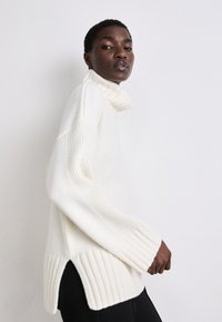 Weißer Pullover mit hohem Kragen, gerippten Bündchen und Saum, versehen mit seitlichen Schlitzen. Weicher Stoff, lockere Passform und minimalistisches Design.