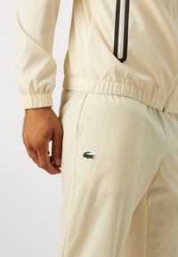 Cream tracksuit made of lightweight fabric. Features elastic cuffs, zip pocket, and a green crocodile logo on the pants. Smooth texture.