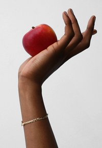 Red apple held in a dark brown hand; gold chain bracelet on wrist. Simple background highlights the apple and hand's texture.