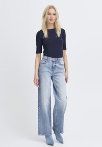 Marineblaues Kurzarmoberteil mit figurbetontem Schnitt; hellblaue, weit geschnittene Jeans mit hoher Taille; das Modell trägt hellblaue Stiefel mit Absatz.