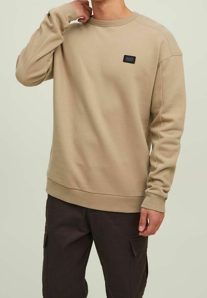 Sweat-shirt beige à col rond avec manches longues et petit écusson logo noir sur la poitrine, porté avec un pantalon cargo foncé.