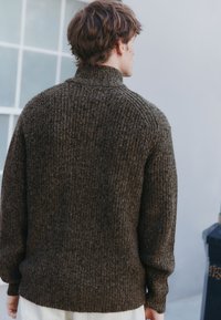 Bruine gebreide trui met een hoge kraag, met een geribbelde textuur en een losse pasvorm, gezien van achteren. Lichtgekleurde broek zichtbaar.