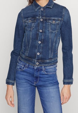 Personne portant une veste en denim bleu ajustée avec fermeture à boutons et un jean en denim bleu assorti sur un fond uni.