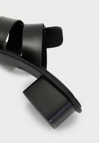 Sandales à talons noirs en cuir lisse, avec deux larges brides et un talon épais. Le design est minimaliste avec une finition mate.