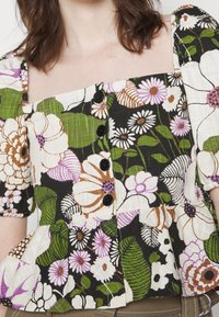 Femme portant un chemisier à motif floral avec des manches bouffantes et des boutons noirs sur le devant, avec des fleurs blanches, vertes, marron et violettes.