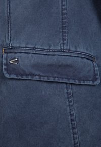 Gros plan d'un tissu en denim bleu montrant un rabat de poche horizontal avec des coutures détaillées et un petit logo métallique sur le côté gauche.