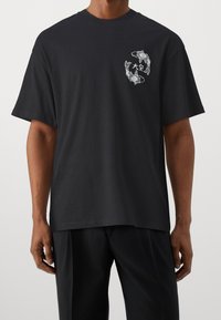 Svart bomullst-shirt med korta ärmar, med en grafisk design av koi-fiskar och stjärnor på vänster bröstområde. Mjuk, avslappnad passform.