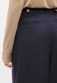 Tengerészkék nadrágok hátsó zsebekkel és arany rózsa alakú gombokkal a derékon, bézs színű, hosszú ujjú felsővel párosítva. Simaság tapintású anyag.