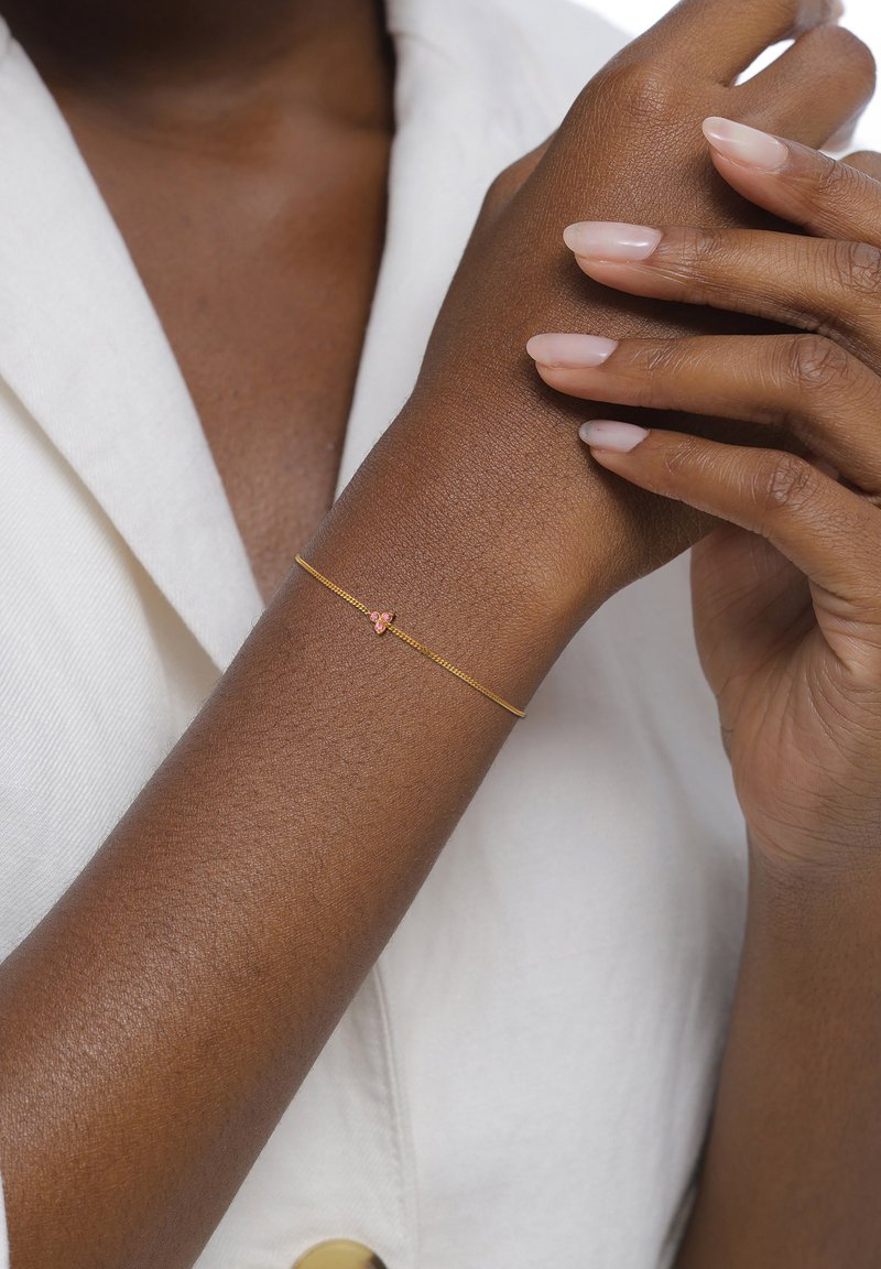 Bracelet en or avec un design de chaîne délicate, comprenant un petit accent en pierre précieuse rose. Le teint de la peau est profond et texturé. Tissu blanc visible.