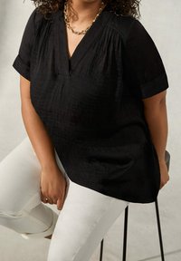 Blusa nera a maniche corte con scollo a V, tessuto testurizzato e dettagli plissettati. Abbinata a jeans bianchi e una collana d'oro.