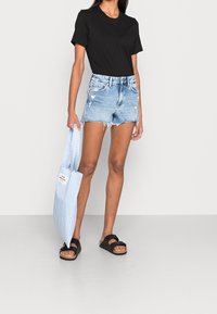 T-shirt noir à manches courtes, shorts en denim bleu clair déchirés, sandales noires, et un sac fourre-tout bleu et blanc rayé avec une étiquette.