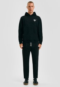 Zwarte hoodie met een voorzak en logo, gecombineerd met bijpassende joggingbroek. Glad materiaal, ontspannen pasvorm en witte trekkoorden. Zwarte sneakers.