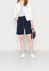 Marine cargoshorts met een strikceintuur, gecombineerd met een wit shirt met stippen en een lichte spijkerjack. Witte sneakers en een kleine zwarte handtas.