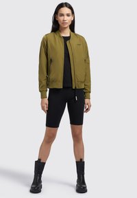 Olive roheline bomber-jakk ribidega varrukatega, lukuga taskud ja lukuga esikülg, koos musta topi ja mustade rattashortsidega.