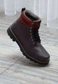 Dark brown leather boot with red padded collar, black laces, rugged sole, and embossed logo on side, resting on marble floor near white curtains.