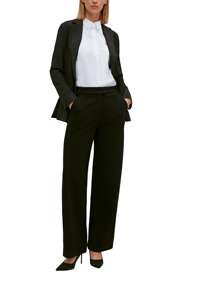 Schwarzer maßgeschneiderter Blazer über einem weißen Hemd mit Knöpfen, kombiniert mit schwarzen Palazzo-Hosen und schwarzen High Heels.