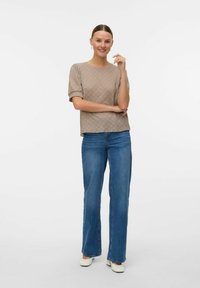Beige texturerad topp med korta puffärmar, parat med blå raka jeans och vita skor. Modellen poserar med en hand på hakan.