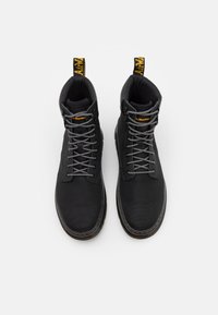 Paire de bottes montantes noires avec des lacets noirs et blancs et un marquage jaune sur la languette et les tirettes talonnières sur fond blanc.