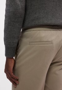Maglione grigio a coste abbinato a pantaloni beige, con tasca posteriore e texture liscia. L'insieme presenta una vestibilità comoda e dettagli minimalisti.
