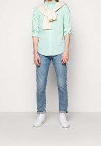 Grön- och vitrandig skjorta med knäppning och långa ärmar, matchad med ljusblå jeans och vita sneakers. En tröja ligger över axlarna.