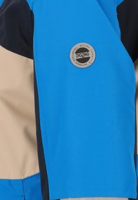 Sininen ja beige takki, jossa on teksturoitu pinta, ja jossa on pyöreä hopeinen logo, jossa on teksti "ZIGZAG". Mukana mustat aksenttipaneelit.