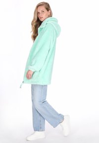 Menta zöld bolyhos kapucnis dzseki cipzáros záródással és állítható húzózsinórokkal, világoskék farmernadrággal és fehér tornacipővel párosítva.