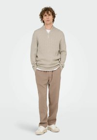 Junger Mann mit lockigem Haar, der einen beigen Pullover mit Kabelstrick und halbem Reißverschluss, taupefarbene Hosen, ein weißes T-Shirt und weiße Sneakers trägt, steht mit den Händen in den Taschen.