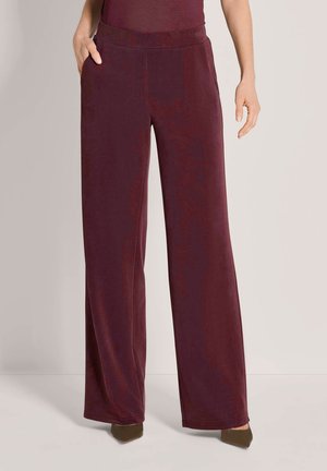 Vrouw draagt wijde bordeauxrode broek met zakken, gecombineerd met donkere puntige schoenen tegen een effen achtergrond.