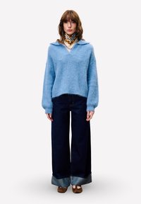 Hellblauer, flauschiger Pullover mit V-Ausschnitt und weiten Ärmeln, kombiniert mit dunkelblauen, weiten Denim-Jeans und braunen Schuhen mit abgerundeten Zehen.
