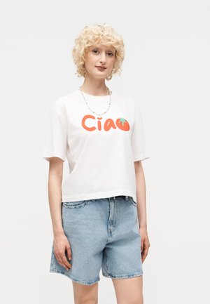 Νεαρό άτομο με σγουρά ξανθά μαλλιά, φορώντας λευκό μπλουζάκι "Ciao" με γραφικό τομάτας και ανοιχτό μπλε τζιν σορτς.